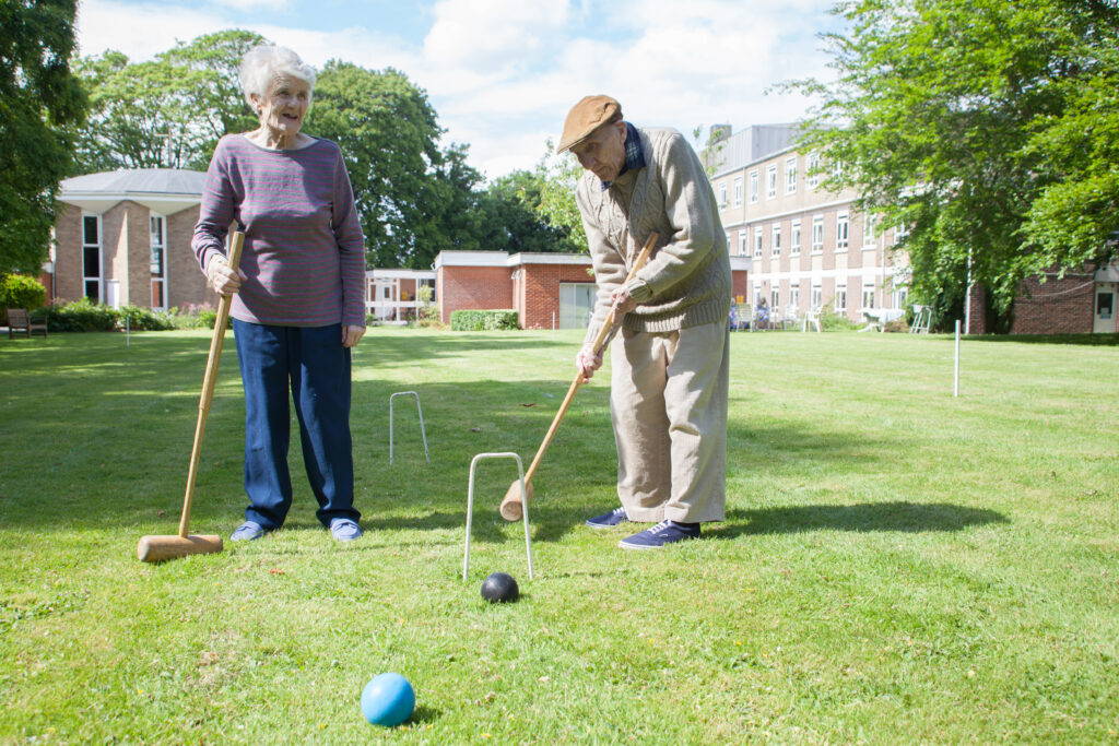 Elderly couple playing croquet on a sunny lawn, surrounded by trees and buildings in the background.