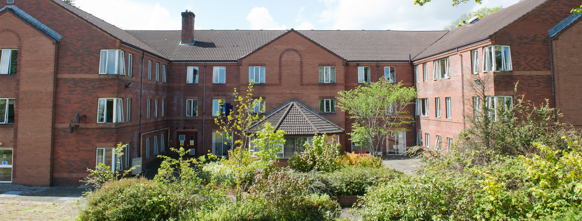 Red-brick care home building with multiple windows, a central gazebo, and landscaped gardens under a clear blue sky.