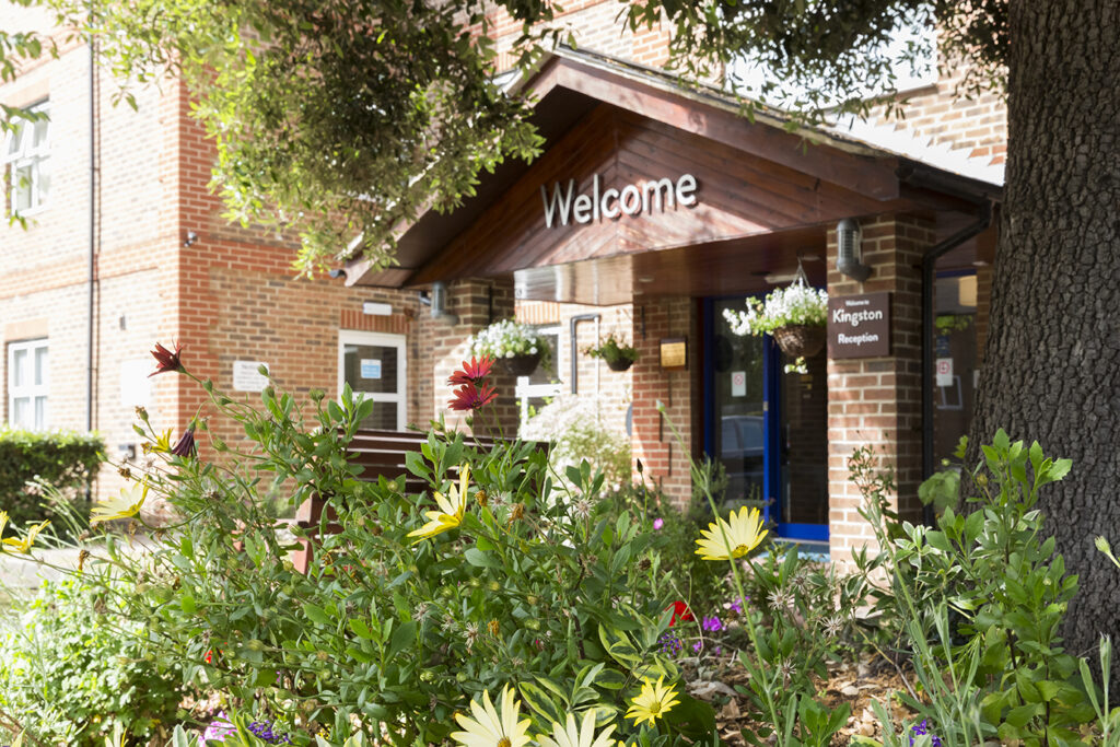 Front entrance of Kingston Care Home with a welcoming sign, surrounded by vibrant flowers and greenery, under a sunny sky.