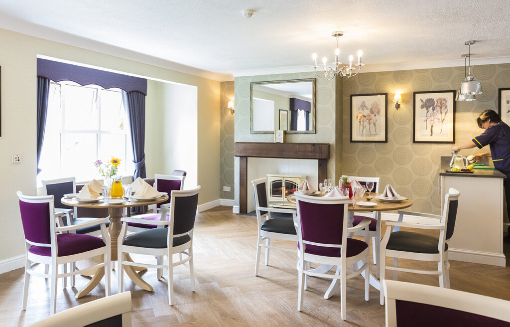 Bright dining area featuring elegant round tables with purple and black chairs, adorned with flowers and table settings, and cozy decor.