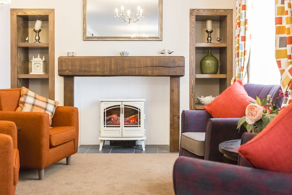 Cozy lounge area featuring orange and patterned chairs, a wooden fireplace with a decorative mantel, and bright curtains.