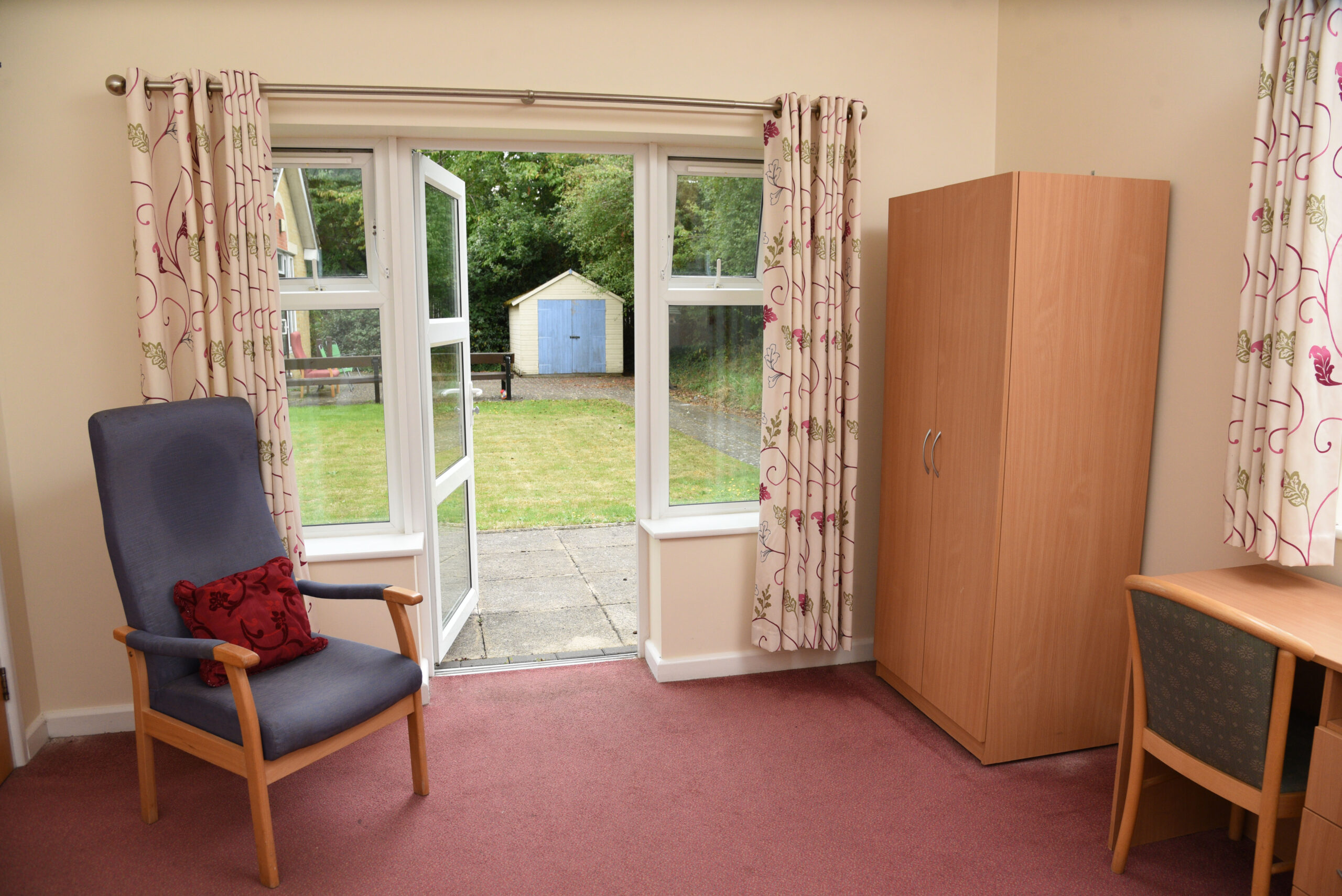 Bright bedroom featuring a blue accent chair with a cushion, wooden wardrobe, and a desk, with patio doors opening to a garden view.