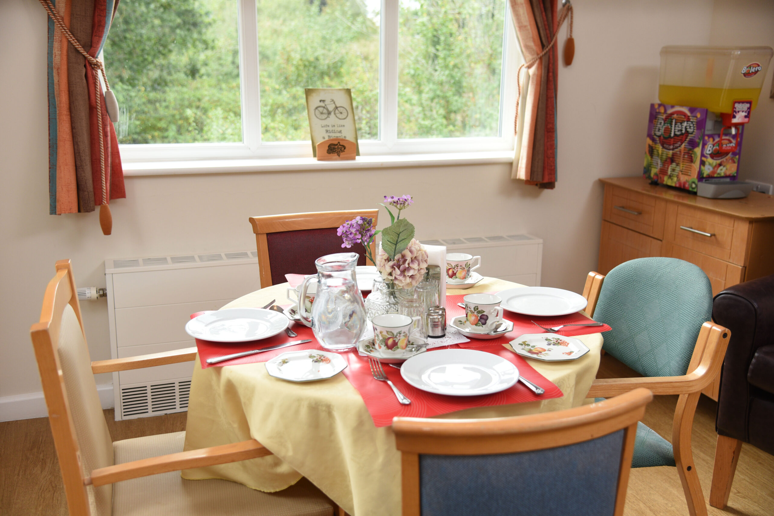 Set dining table with floral decor, dishes, and glassware, surrounded by colorful curtains and cozy furniture, overlooking greenery.