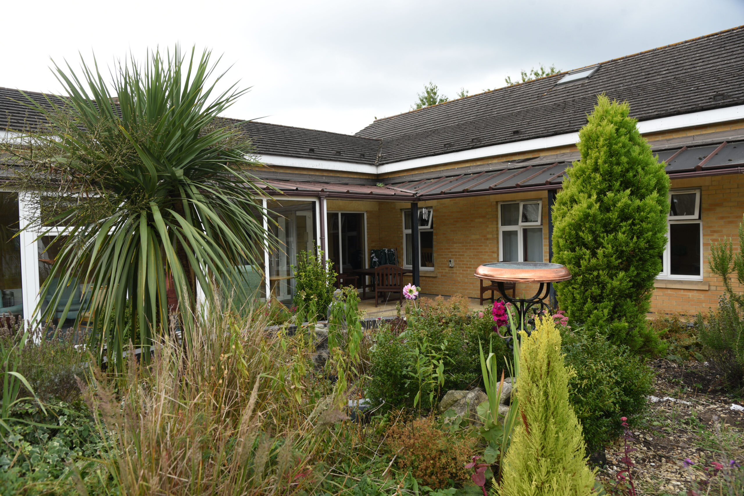 Lush garden featuring a variety of plants, a small table, and seating areas by a brick building with overhanging eaves.