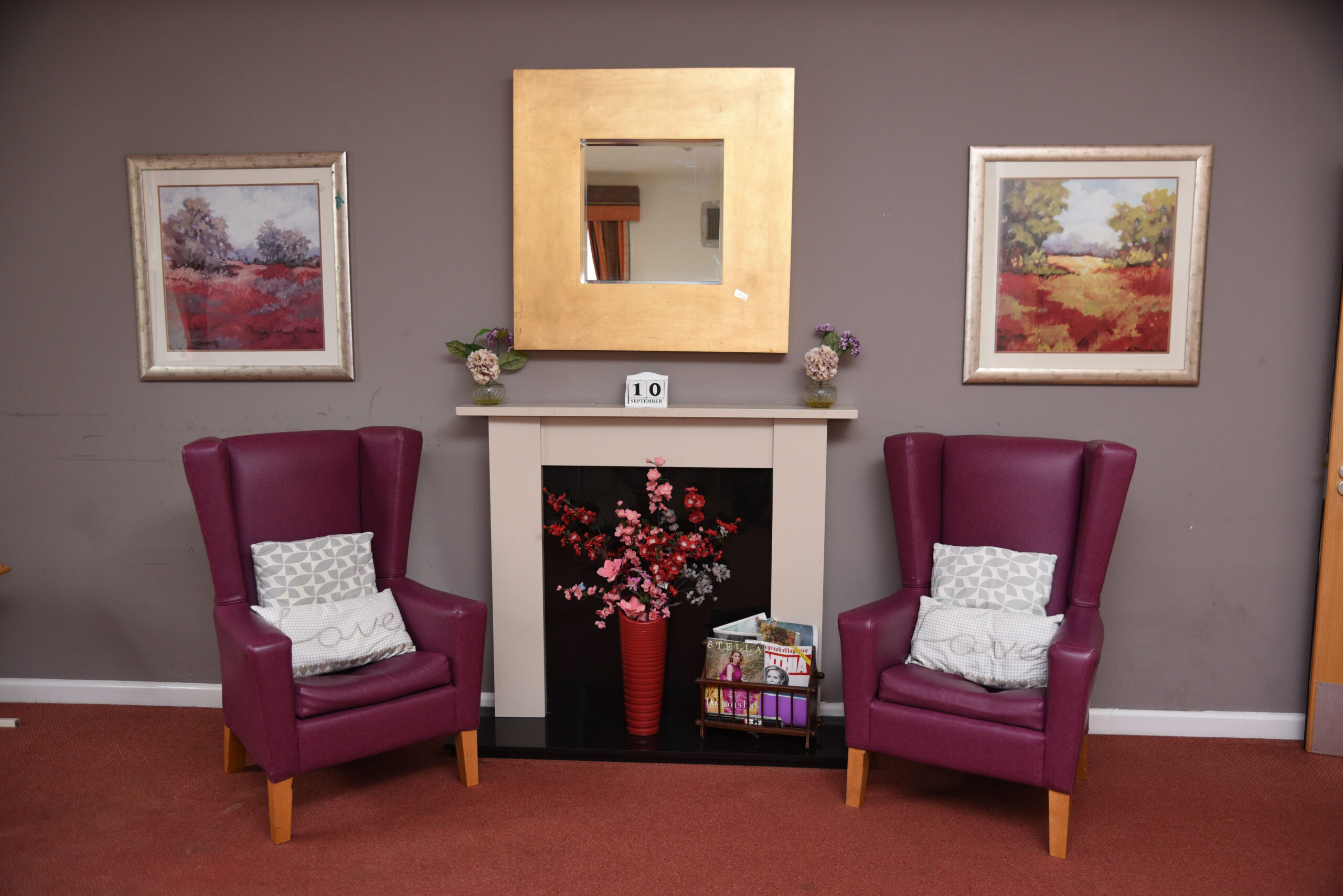 Cozy lounge with two purple armchairs flanking a fireplace, adorned with a mirror and paintings, creating a welcoming atmosphere.