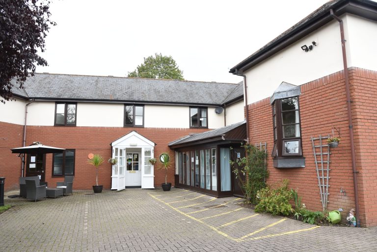 Exterior view of The Triangle Care Home featuring a brick facade, entrance with white doors, landscaped areas, and seating, a care home in Wheatley.