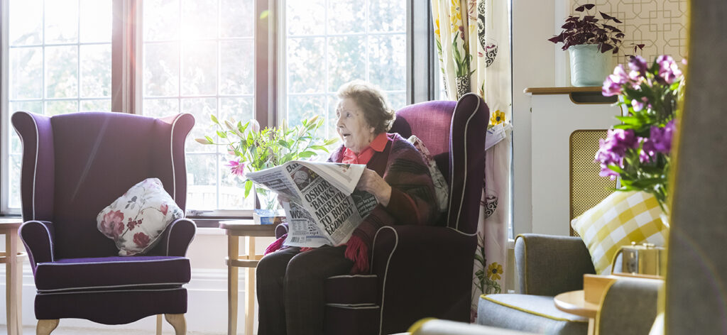 A resident reading a newspaper in a cozy, well-lit lounge, surrounded by plants and flowers in a welcoming care home setting.