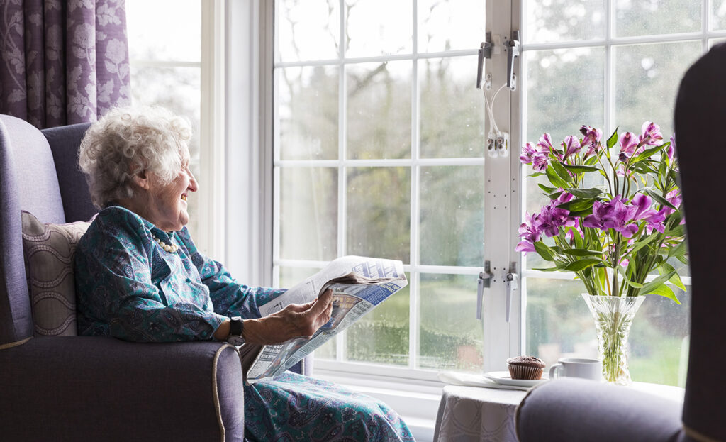 An elderly woman smiles while reading a newspaper by a window, with a vase of purple flowers and a cupcake on the table beside her.