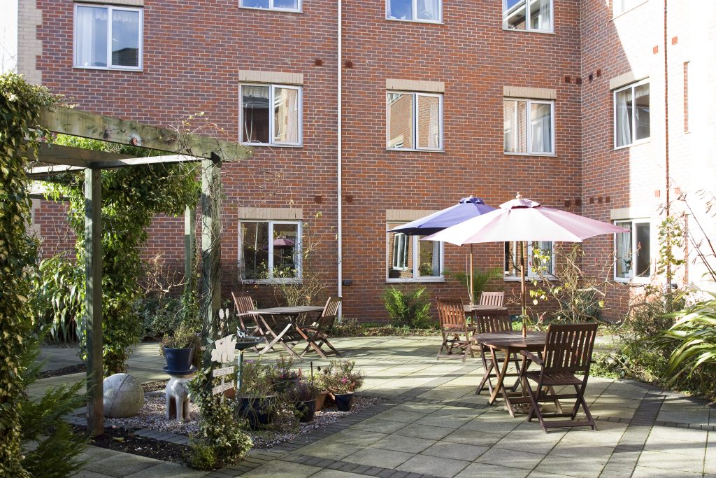 Serene courtyard at Lansdowne Care Home featuring wooden tables and chairs, a pergola, and colorful umbrellas, surrounded by greenery.