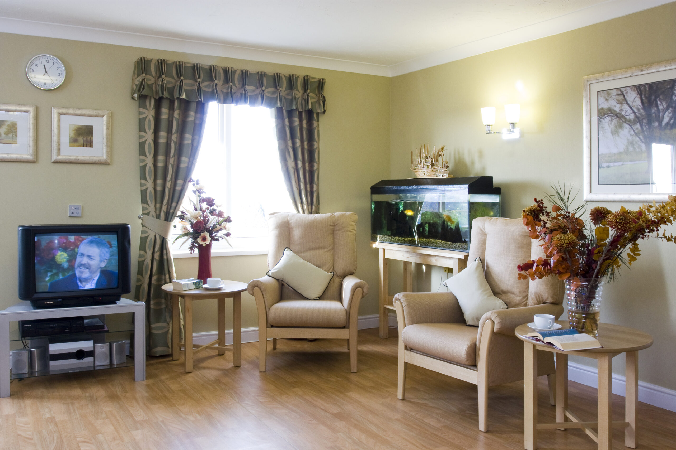 Cozy lounge area with two cream armchairs, a coffee table, an aquarium, a TV, and decorative flowers, featuring warm, inviting colors.
