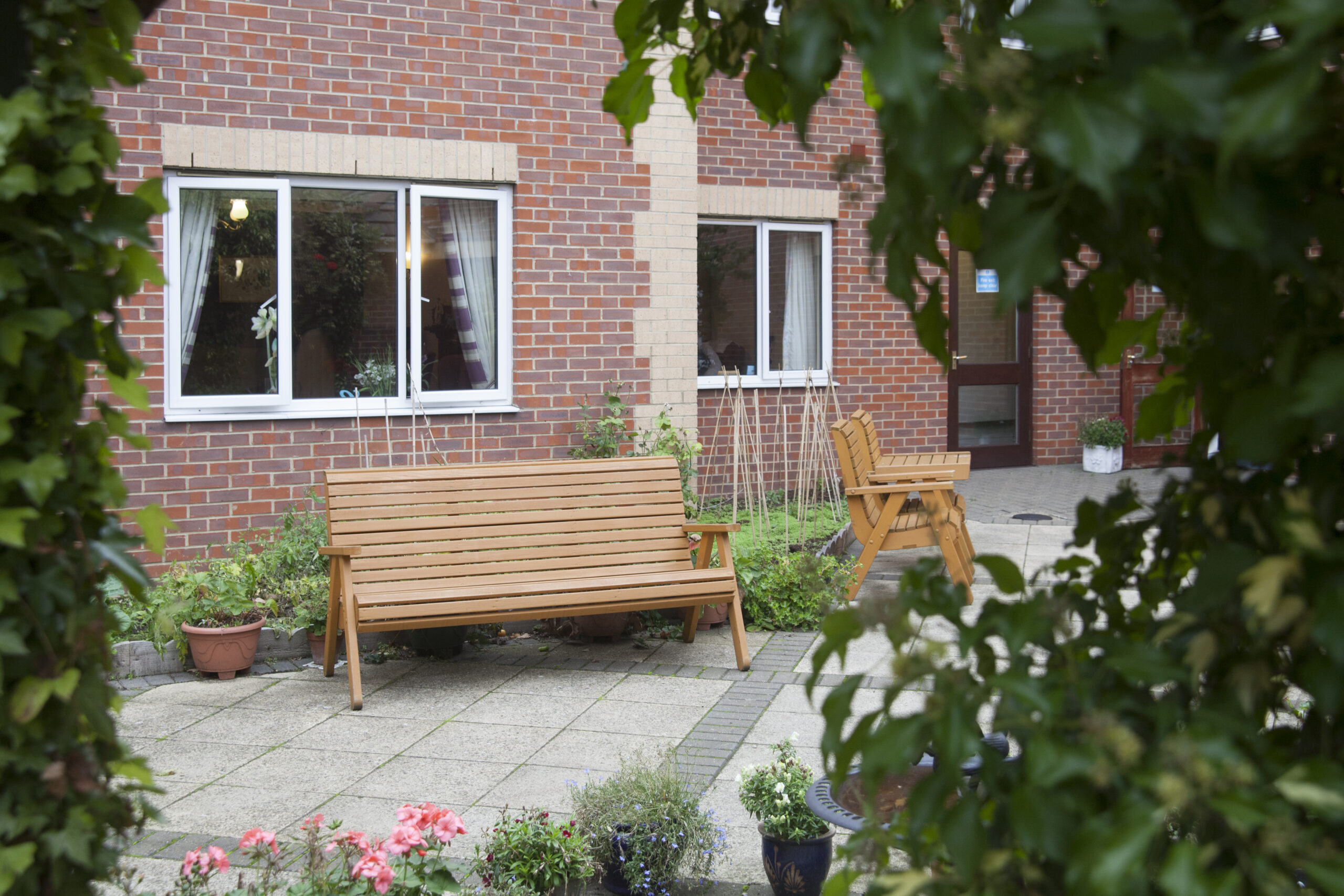 Wooden benches arranged in a courtyard surrounded by greenery and flowers, with brick walls and windows of the Lansdowne Care Home in view.