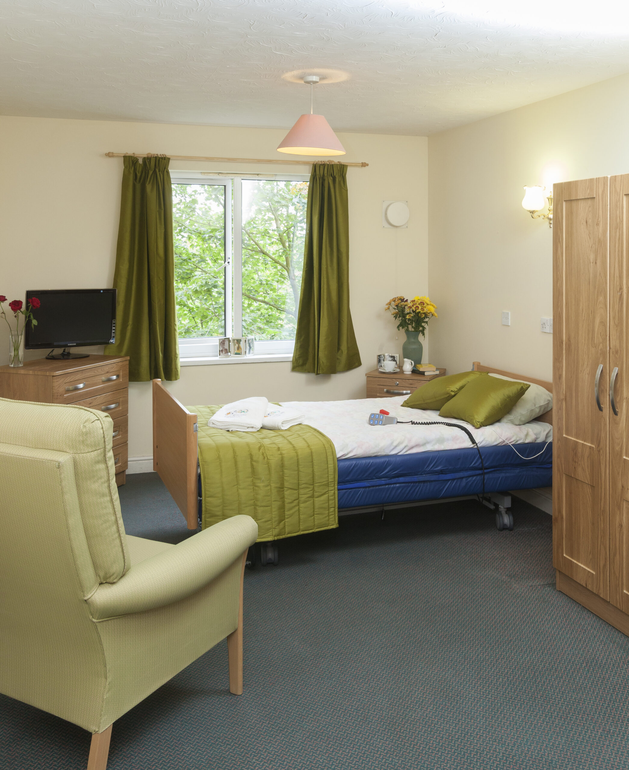 Bright, welcoming room in Lansdowne Care Home featuring a single bed, green curtains, a TV, and a cozy armchair by the window.