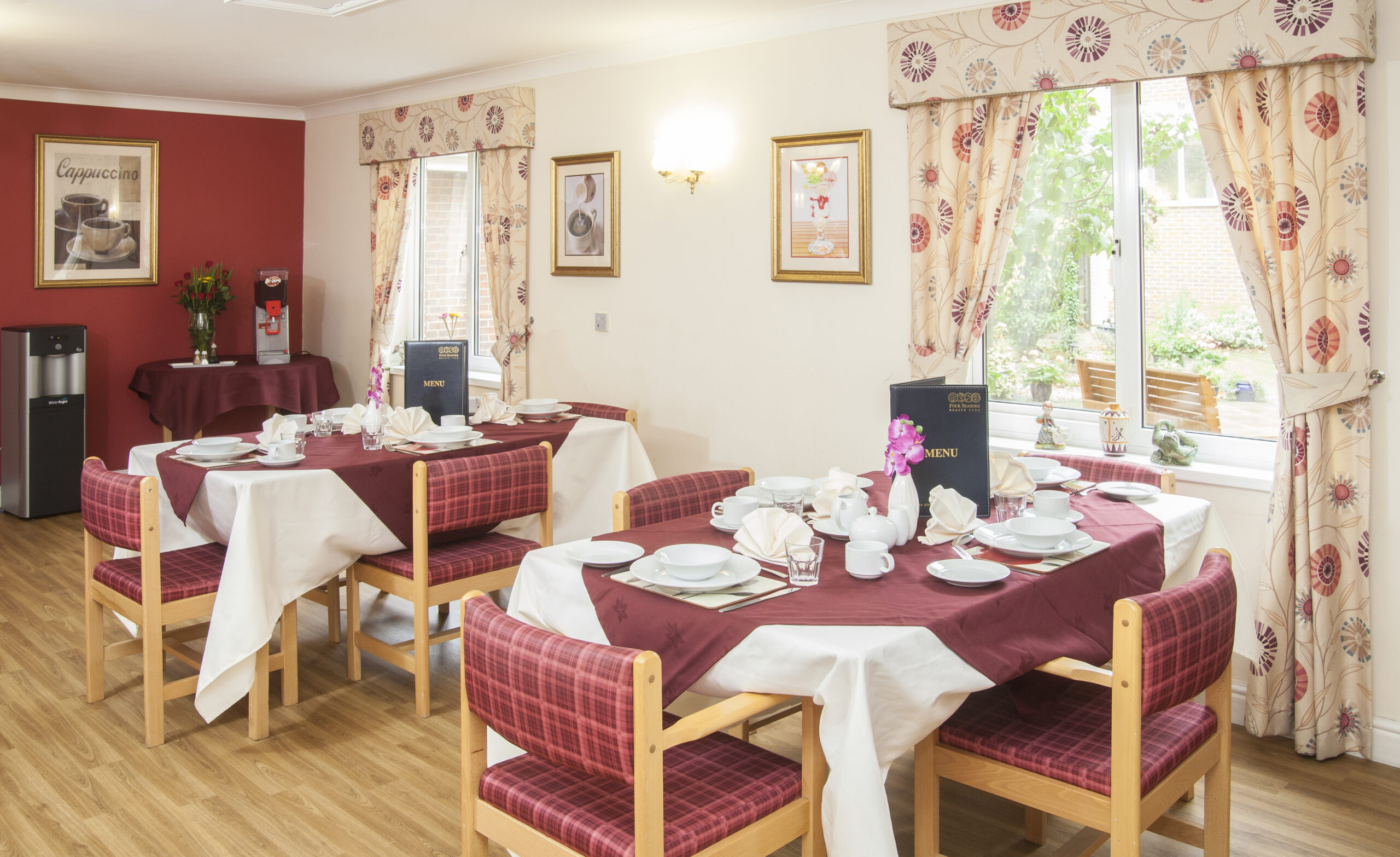 Elegant dining area in Lansdowne Care Home featuring tables set with white tableware, maroon accents, and floral curtains by a sunny window.