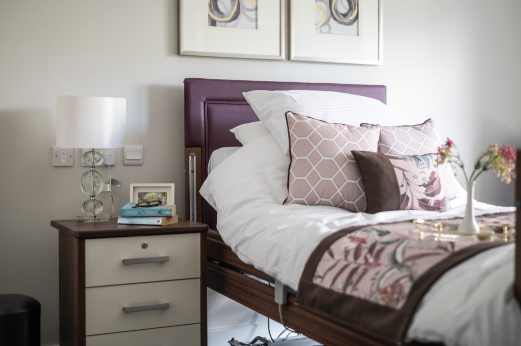A furnished bedroom with a purple headboard, bedside table, lamp, books and flowers.