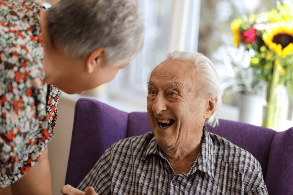 A smiling elderly man sits in a purple chair, laughing with a woman in a floral top, bright flowers in the background.