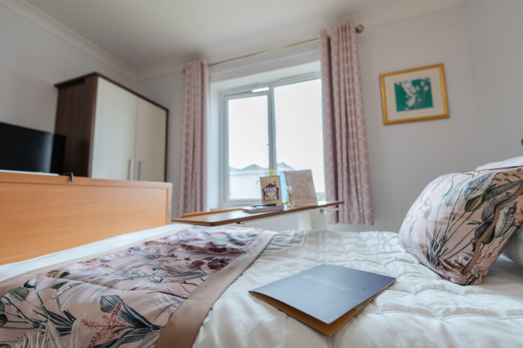 Cosy, well-appointed bedroom at a Willesden care home, featuring a floral bedspread, bedside table with framed photos and soft pink curtains.