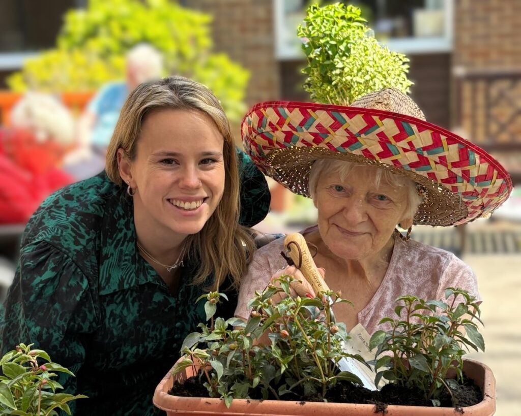 A woman and an older woman pose with potted plants at Queensway House; the older woman wears a colourful woven sun hat.