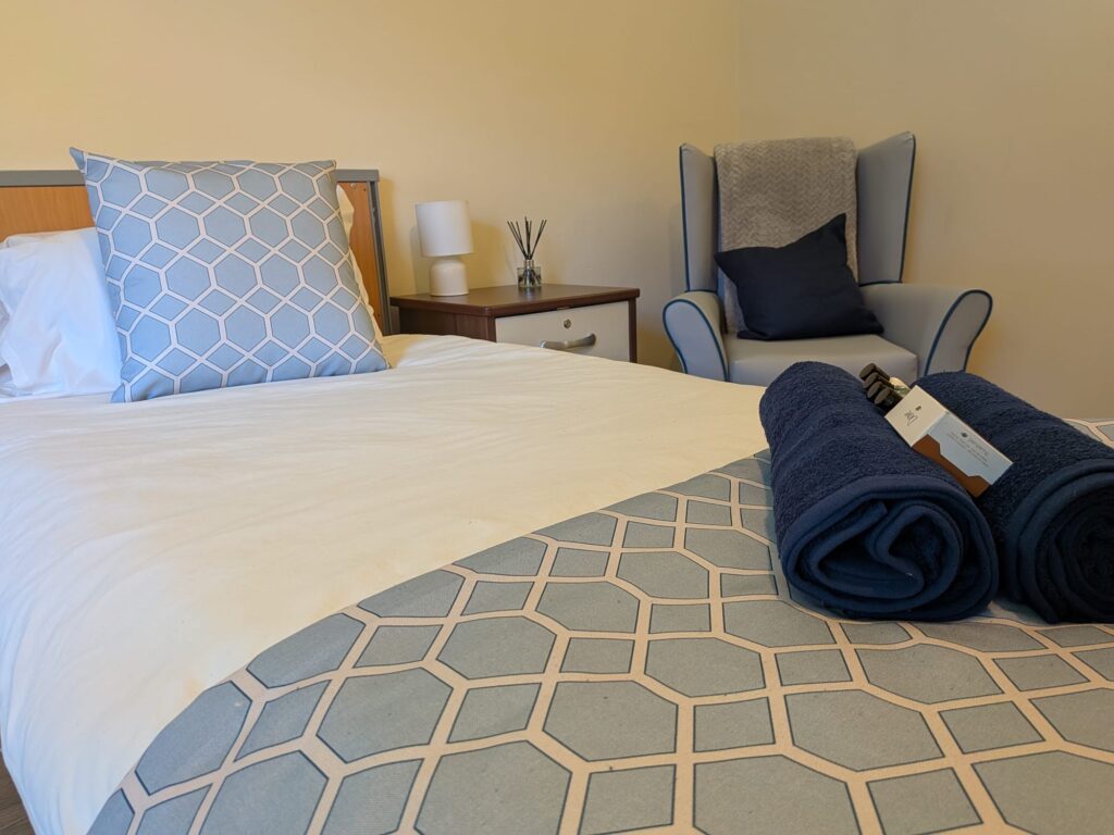 Queensway House bedroom with a made bed, folded towels, an armchair and a bedside table.