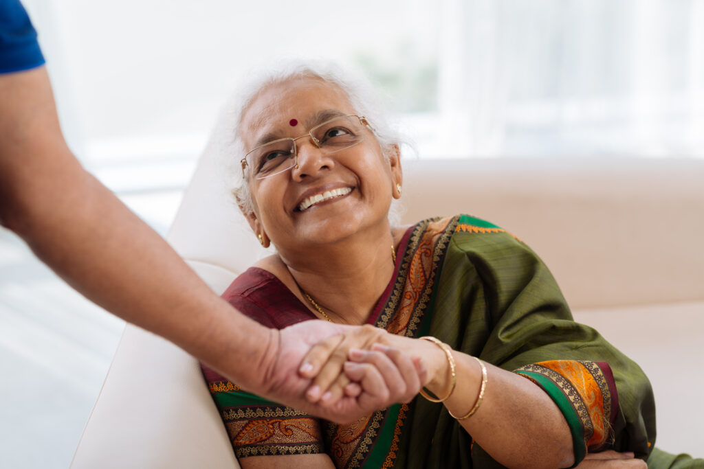 An elderly woman in a colorful sari smiles warmly as she holds hands with a caregiver, seated in a bright, welcoming space.