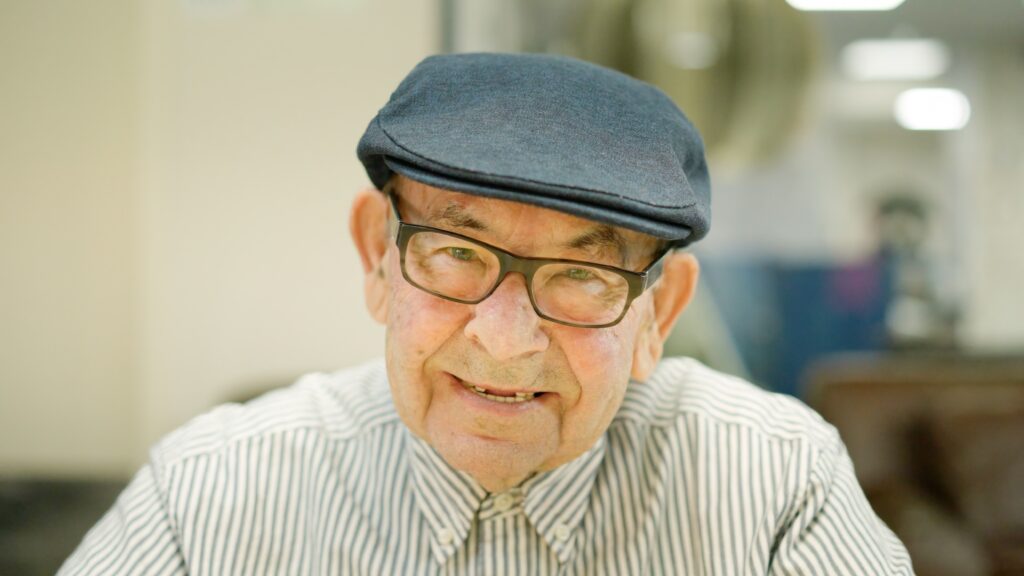 An elderly man with glasses and a navy cap smiles warmly, dressed in a striped shirt, against a softly blurred background.