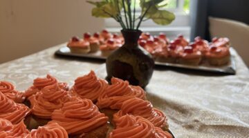 A tray of orange frosted cupcakes in the foreground, with a vase of flowers and more cupcakes blurred in the background by a window.