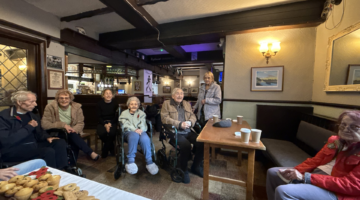 Residents and staff gather in a cozy pub setting for Remembrance Day, with snacks on a table and poppies worn by attendees.
