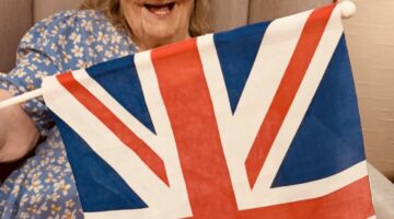 Elderly woman in a floral dress happily holding a Union Jack flag while seated in a cozy armchair, smiling in a warm setting.