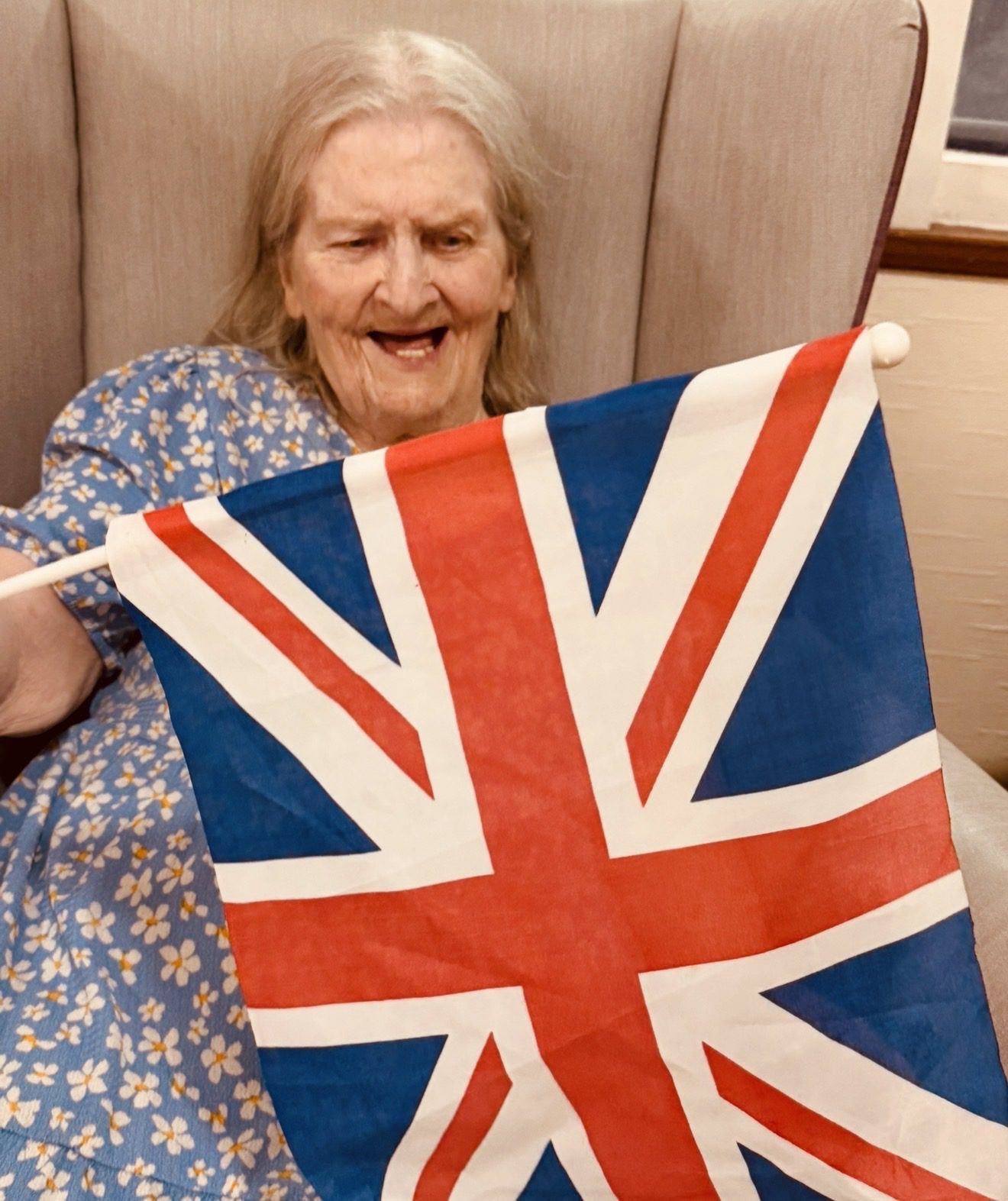 Elderly woman in a floral dress happily holding a Union Jack flag while seated in a cozy armchair, smiling in a warm setting.
