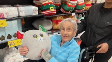 Elderly woman in a wheelchair holding a plush Christmas-themed pillow, smiling, with festive decorations on store shelves behind her.
