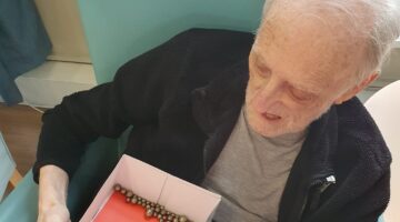Elderly resident seated in a light blue armchair, engaging with a sensory activity box featuring a teddy bear design and wooden beads.